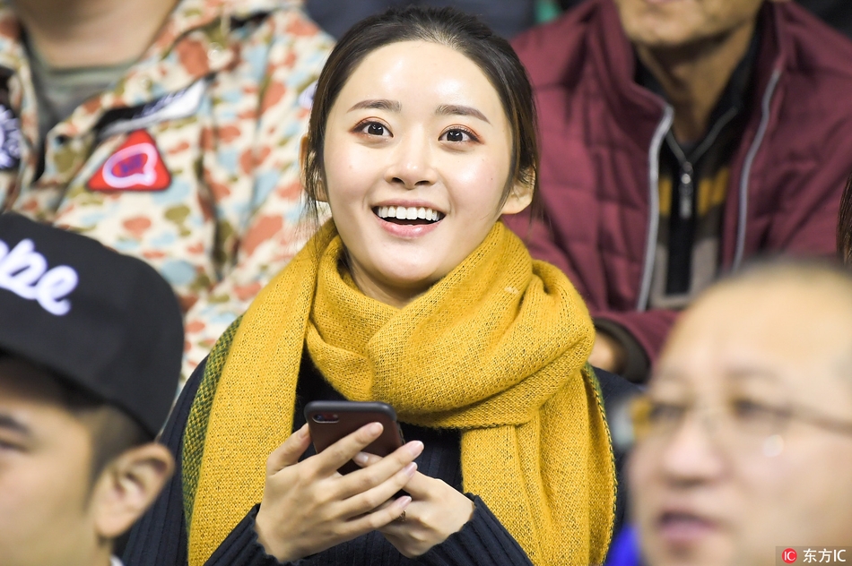 一名身穿时尚休闲装的女孩在看台上回眸，背景是欢呼雀跃的人群，她的嘴角带着淡淡的微笑，显得既冷静又迷人。