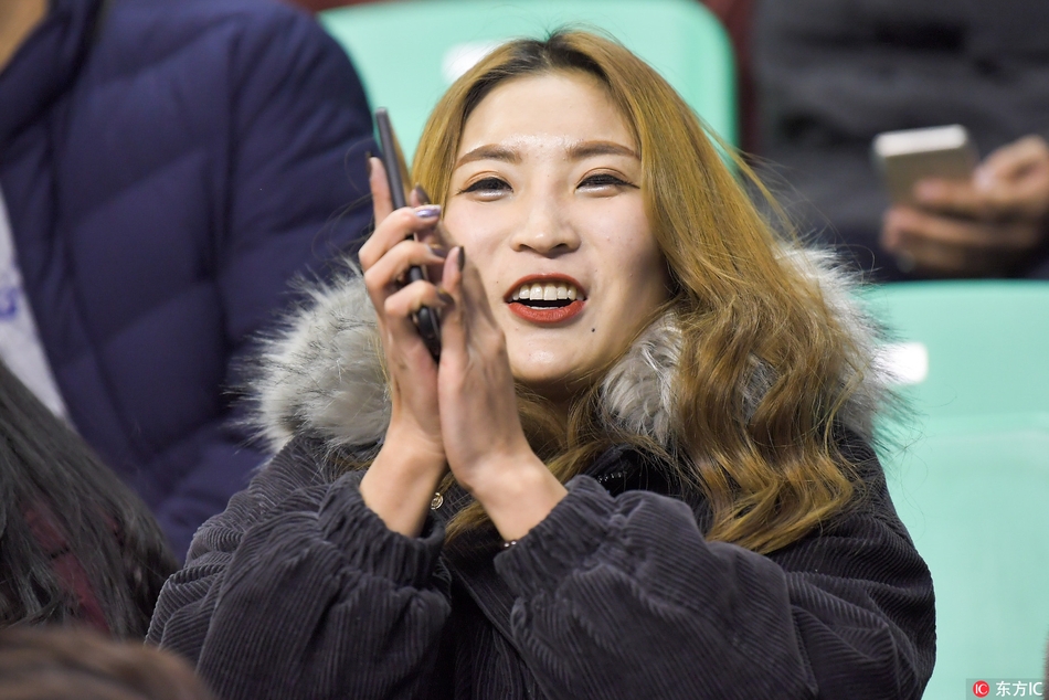 一位长发披肩的优雅女球迷，面庞在体育馆灯光的映照下显得格外立体，她目光如炬，仿佛在审视场上每一个战术变化。