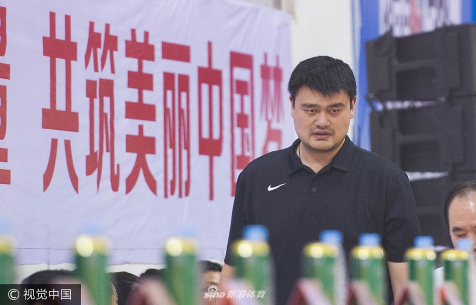 在喧闹的体育馆内，姚明侧身倾听身旁友人的话语，表情认真而专注，偶尔点头回应，眼神中透露出对场上局势的关切与思考。