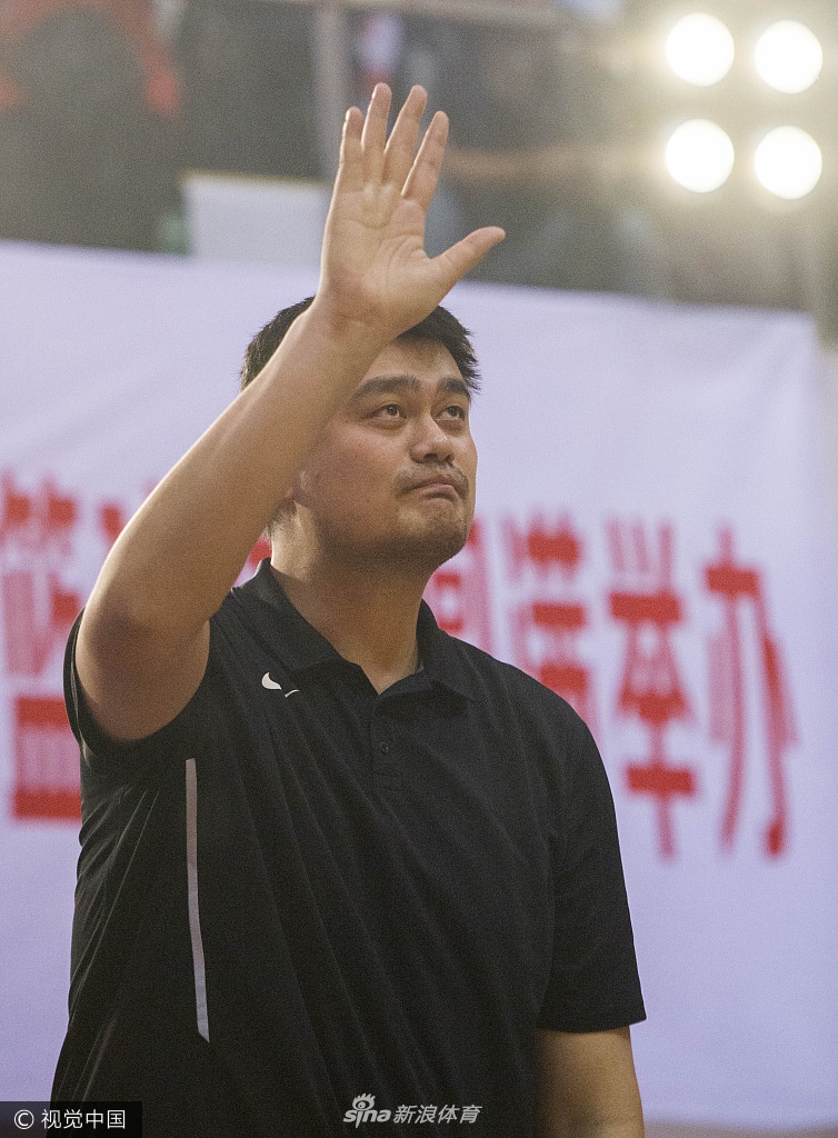 姚明站立在人群中，他的身高让他显得格外突出。他神情专注地看着场内的比赛，脸上带着一丝严肃，仿佛在思考着中国篮球基层联赛的下一步发展方向。