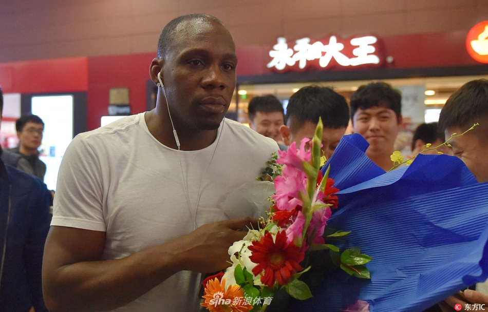 哈德森手推行李车走出通道,目光坚定地望向远方,身旁簇拥着满脸笑容的球迷,背景里隐约可见“欢迎回家”的横幅,氛围热烈而有序。