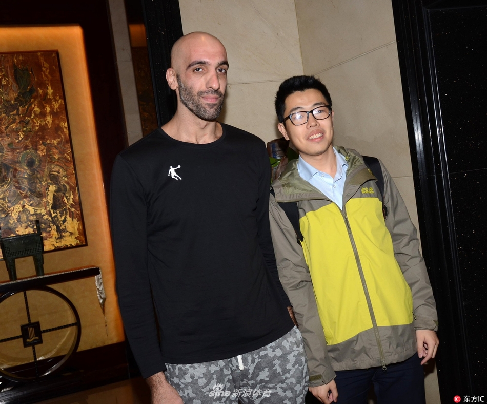 马布里蹲在地上耐心地为几名小球迷签名，脸上带着慈父般的微笑，行李箱随意地倒在脚边，背景是酒店旋转门的暖黄色灯光，定格了这温馨的追星时刻。