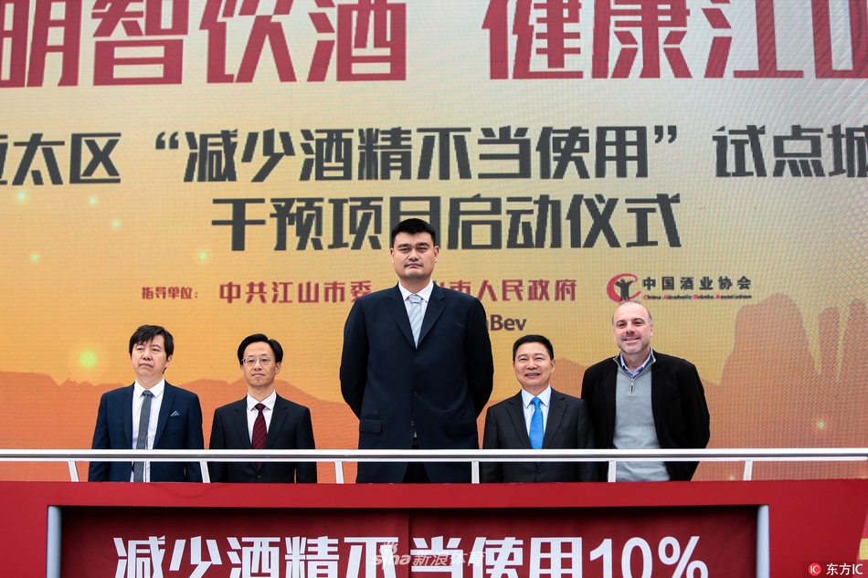 一位身穿黄色公益T恤的工作人员正在为同学们讲解展板上的酒精知识，背景是醒目的“明智饮酒 健康江山”活动标识，姚明站在一旁，认真地观察着同学们的反应。