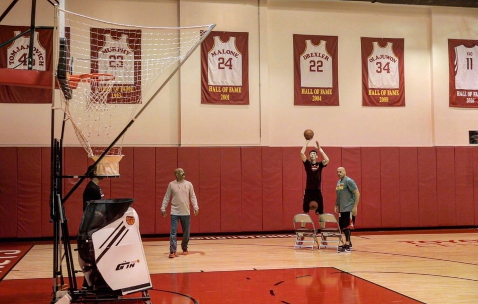 训练场上的周琦神情专注，正在完成一次投篮练习。阳光透过球馆的窗户洒在他身上，仿佛为他镀上了一层金色的光芒。从CBA到NBA，从新疆到休斯敦，他正脚踏实地，向着心中的篮球殿堂一步步迈进。