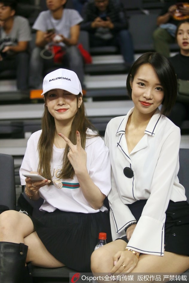 一位气质温婉的女球迷坐在看台前排,背景是激烈的比赛画面,她仿佛沉浸在自己的篮球世界中,温润如玉。