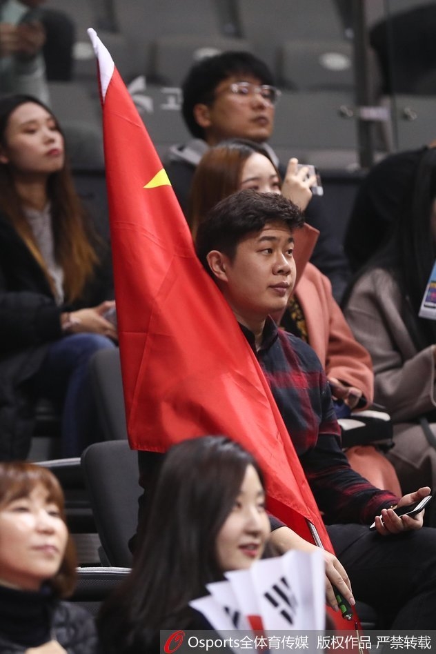 几位中国球迷并肩站立,其中一人高举着五星红旗,他们的目光紧紧锁定在赛场中央。周围的韩国观众神情各异,红色的旗帜在这一片陌生的色彩中显得醒目而坚定。