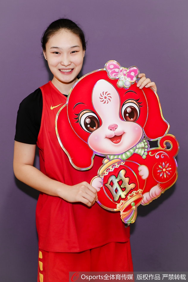 队员们围坐在一起，手中拿着红包和春联，背景的红色与中国女篮的队服交相辉映，画面充满了温馨的大家庭氛围，每个人的脸上都写着对未来的憧憬。