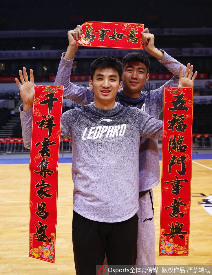 深圳男篮队长孟铎在喜庆的红色背景下，双手紧握鼓槌奋力敲响铜锣，眼神中充满坚定与笑意，仿佛在为即将到来的新春敲开胜利之门。