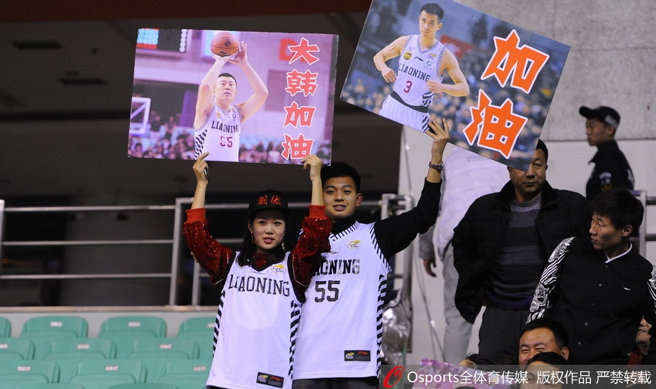 看台上一对父子球迷，父亲满脸笑容地抱着年幼的儿子，儿子手中也拿着助威棒。身后无数双手臂高举，人声鼎沸，父子俩共同享受着篮球带来的快乐时光，这正是球迷文化代代相传的最好写照。