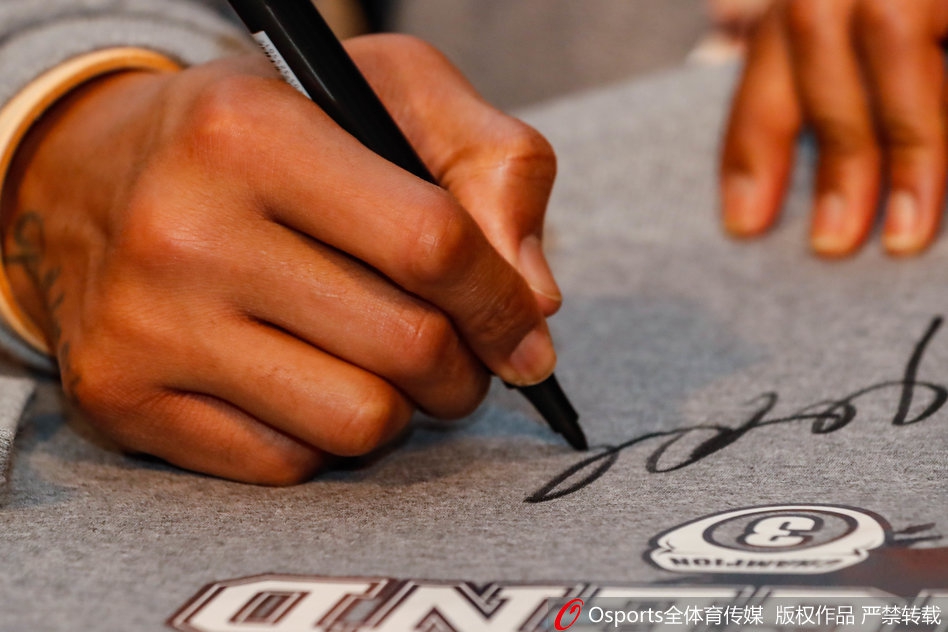 马布里为球迷签名的特写，他手中的笔正在一件卫衣上划过，脸上带着专注而满足的微笑，旁边摆放着球迷精心准备的鲜花和礼物，每一样礼物都承载着球迷沉甸甸的爱。