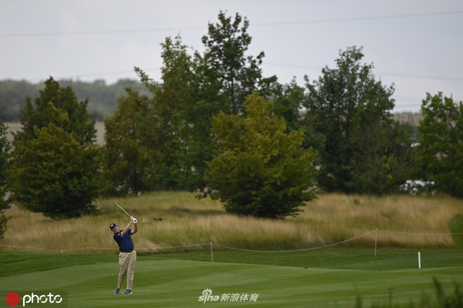 厄尼·埃尔斯在发球台上完成标志性的大幅转体挥杆，球杆在阳光下划出一道银色的弧线，身后是郁郁葱葱的森林球道。
