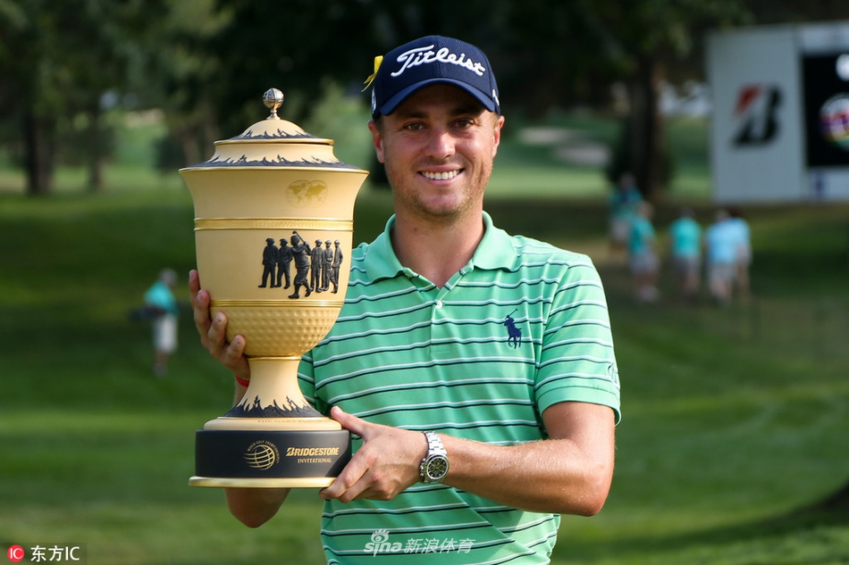 贾斯汀·托马斯在火石乡村俱乐部第十八洞果岭,带着标志性的自信微笑高举普利司通邀请赛冠军奖杯,身后是见证历史的观众与夕阳下的球场。