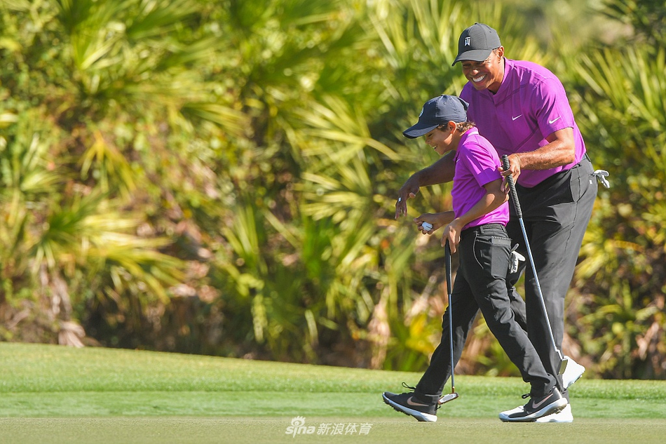 伍兹父子在球道上漫步交流，查理侧耳倾听父亲的指导，背景是优美的球场景观。