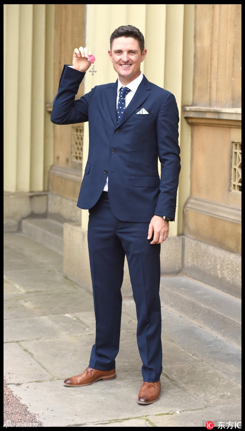 贾斯汀·罗斯面带谦和而自豪的微笑,向镜头展示他刚刚获颁的大英帝国勋章,勋章在光线下熠熠生辉,象征着他辉煌的职业生涯与国家荣誉。