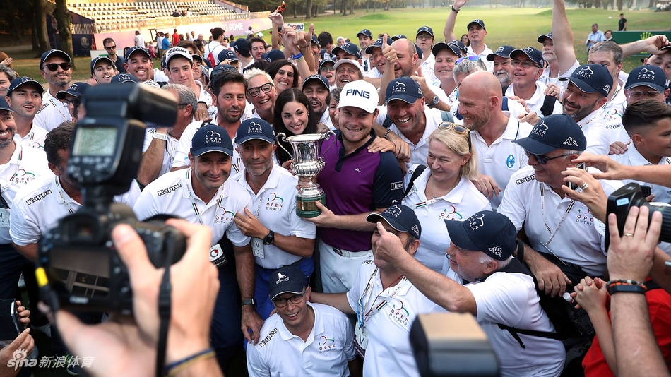 哈顿高举冠军奖杯向观众致意，背景是夕阳下的赛场，金光洒满全身，宛如胜利之神。