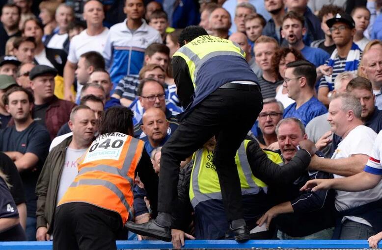 飞出的眼镜在半空中定格，镜片反射着球场刺眼的灯光，背景是模糊不清的看台座椅和其他因震惊而站起的球迷身影。