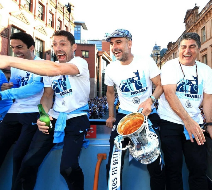 瓜迪奥拉与球员们围成一圈，共同跳跃欢呼，背景是湛蓝的天空和曼城的队旗，展现了这支王者之师无比的团结与活力。