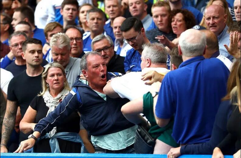 两人扭打在一起，身体失去平衡，手臂纠缠不清，脸部因发力而扭曲变形，蓝白色的球迷围巾在拉扯中显得凌乱不堪。