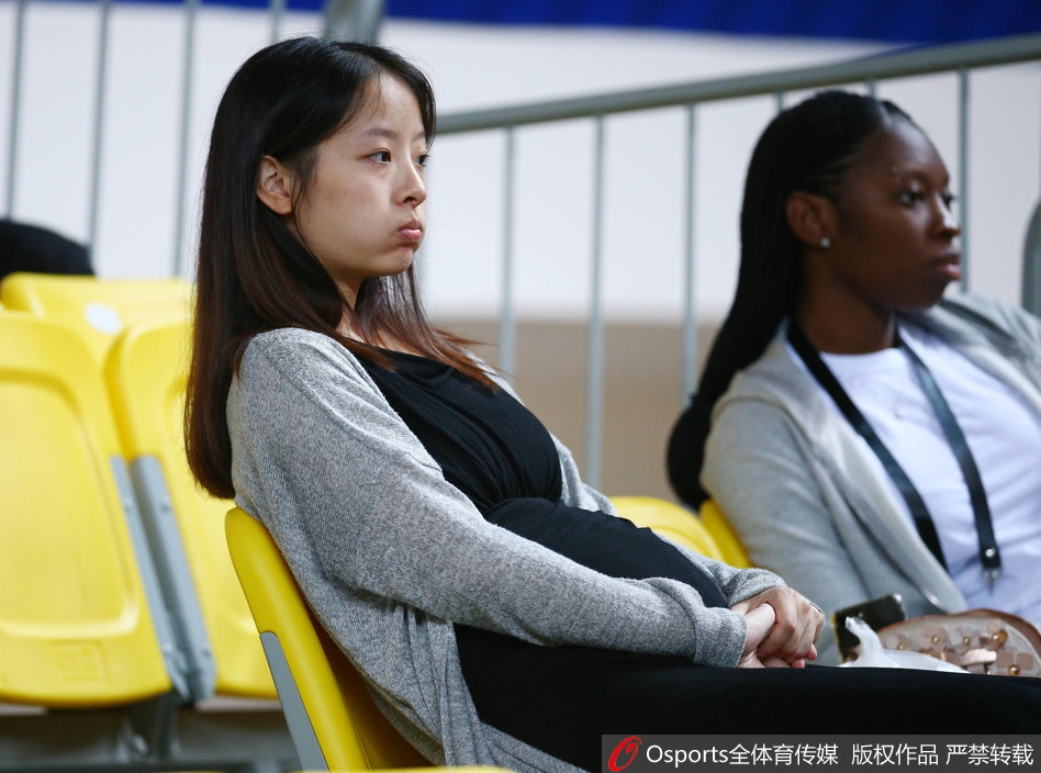 孙桐林妻子身着宽松衣物,双手轻抚在小腹前,眼神温柔地望向赛场方向,脸上洋溢着幸福而含蓄的微笑。