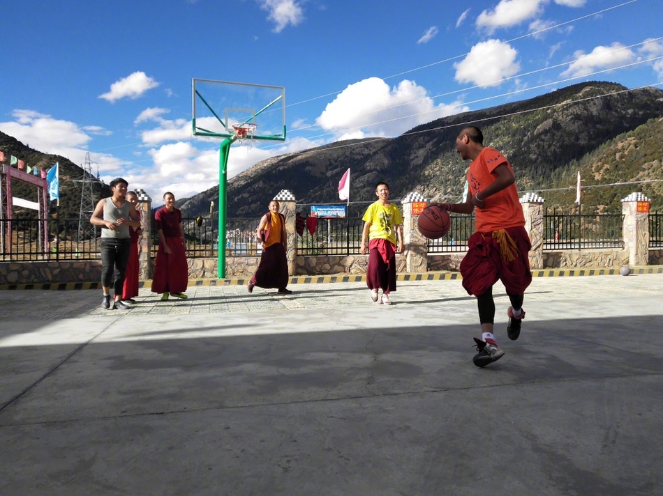 一名年轻的喇嘛正准备投篮,他双腿微曲,双手持球举过头顶,僧袍的衣摆垂落在地。在他身后,是连绵的雪山和湛蓝的天空,构成一幅动静结合、充满禅意的画面。