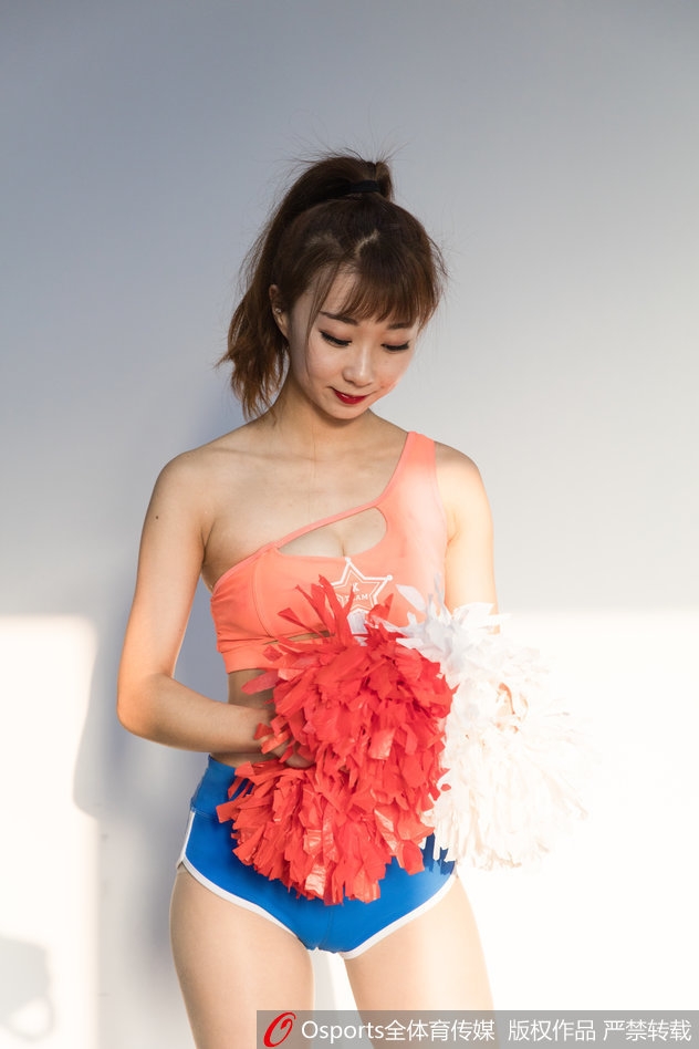 啦啦队女孩扎着高马尾，身穿紫色运动抹胸和白色热裤，手持金色花球，眼神自信地看向镜头