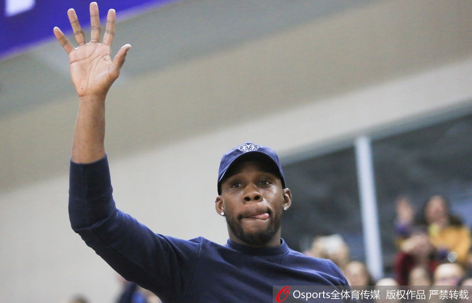 哈德森与身旁的友人交谈，脸上带着轻松的笑容，背景是电子记分牌上辽宁队领先的比分，现场热烈的氛围感染了这位篮球明星。