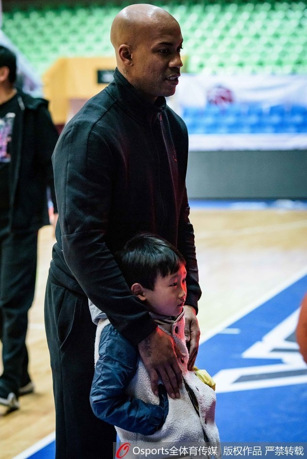 马布里双手抱胸与一位扎着马尾辫的小女孩站在一起，小女孩略显羞涩地比划着爱心手势，老马则低头注视着她，画面定格在刚硬与童真的温柔碰撞瞬间。