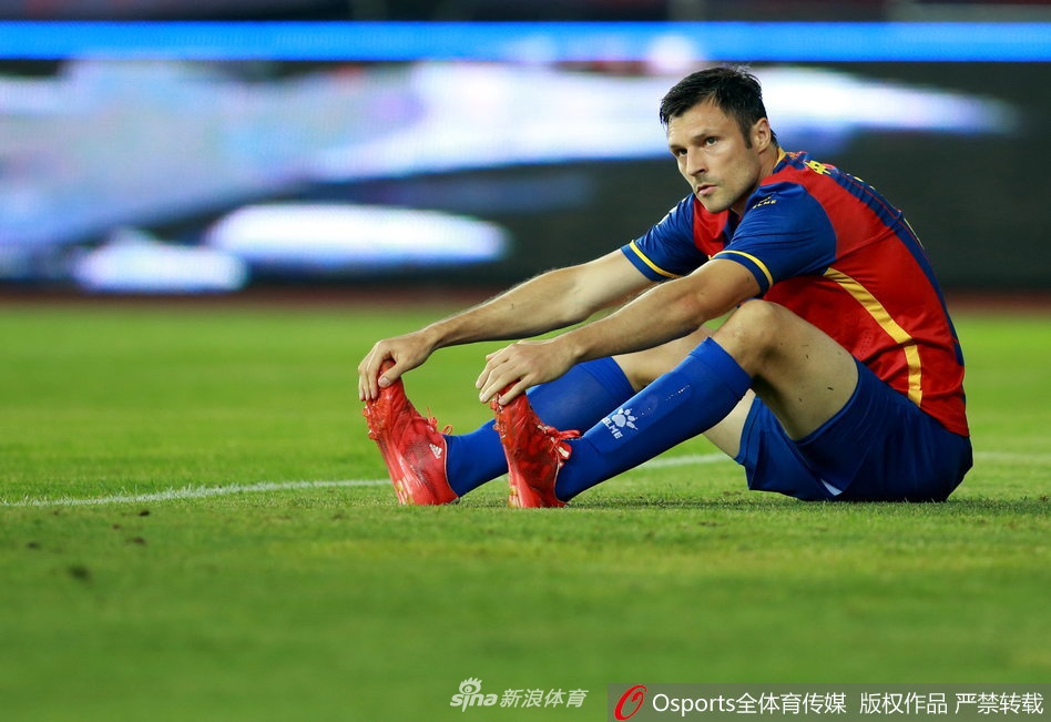 中甲锋霸拉基奇在破门得分后激情四溢地张开双臂庆祝，身后的队友纷纷冲向这位英雄，看台上是挥舞着旗帜、陷入沸腾的青岛球迷，整个球场被橙色的欢乐海洋包围。