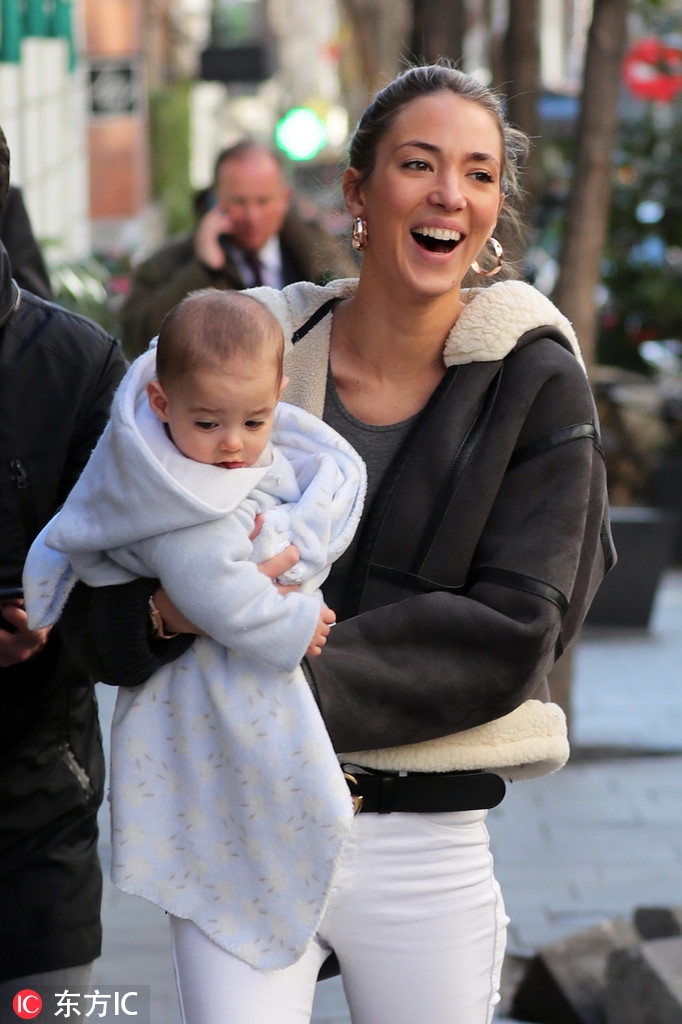 爱丽丝身穿休闲装与莫拉塔并肩行走，她侧身望向丈夫，两人面带微笑，婴儿车中的孩子露出好奇的小脑袋。