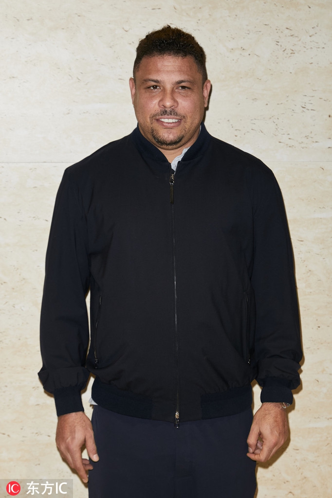 罗纳尔多双手插袋站在背景板前，标志性的笑容让他看起来十分憨厚可爱。