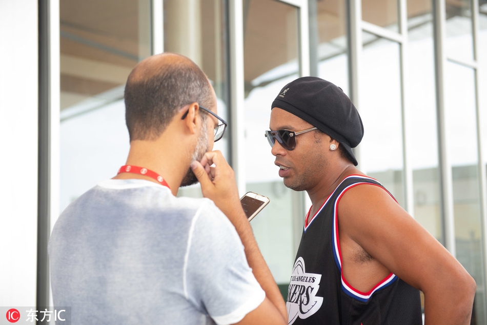 小罗秀出招牌的‘龅牙微笑’，双手指向天空，湖人球衣上的号码在阳光下格外醒目，背后是里约狂欢节五彩斑斓的装饰和兴奋的人群，定格了他回归故土的快乐时刻