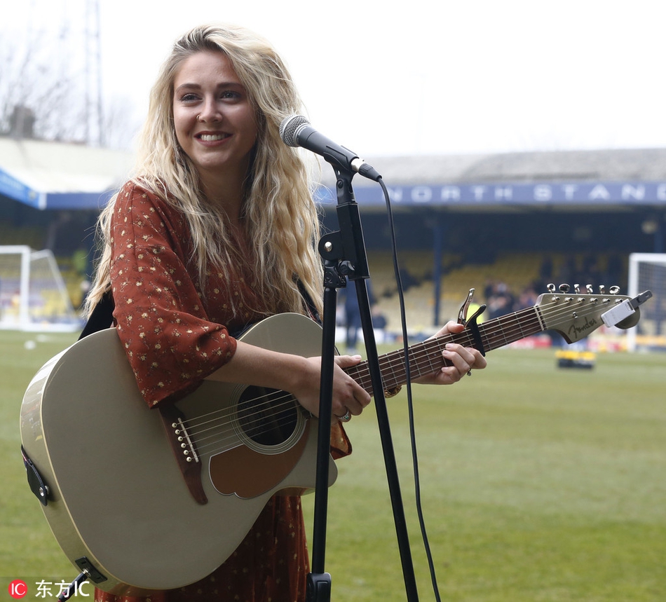 身着时尚便装的梅根-罗斯手持麦克风，在绿茵场上深情献唱，身后是巨大的球场顶棚和灯光，舞台感十足