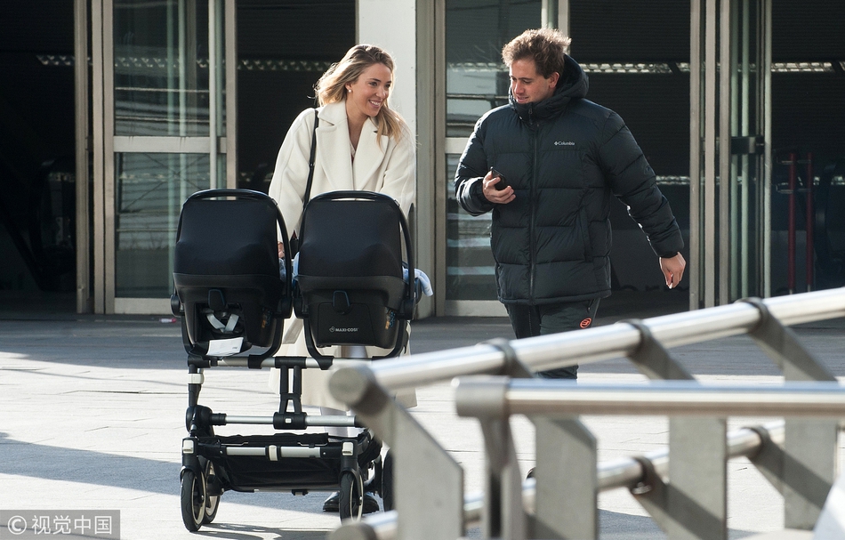 爱丽丝身穿休闲装，推着婴儿车走在马德里繁华的商业区。她高挑的背影与城市的街景交融，步伐坚定，展现了现代母亲兼顾家庭与自我的独立形象。