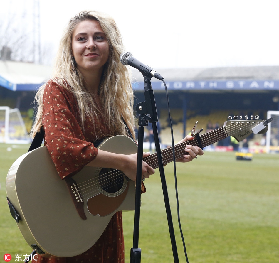 梅根-罗斯在英甲赛场边绽放灿烂笑容，向看台球迷挥手致意，背景是充满英伦风情的球场看台和广告牌
