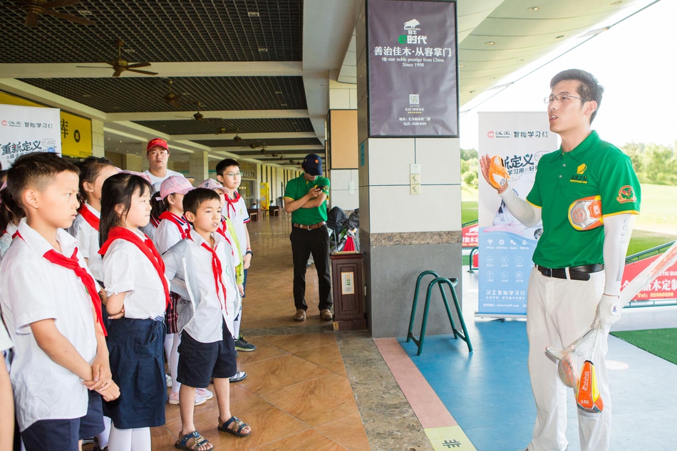 一群戴着太阳帽的小学生,在志愿者老师的带领下,排着整齐的队伍参观球场,他们仰头张望,对广阔的球场和远处挥杆的球员充满了好奇