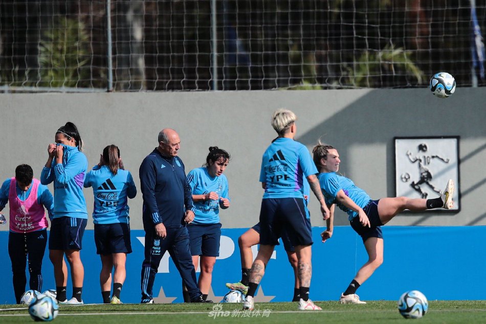 两位女足队员并肩而坐系鞋带，专注的神情与身后辽阔的训练场形成静与动的诗意对比。