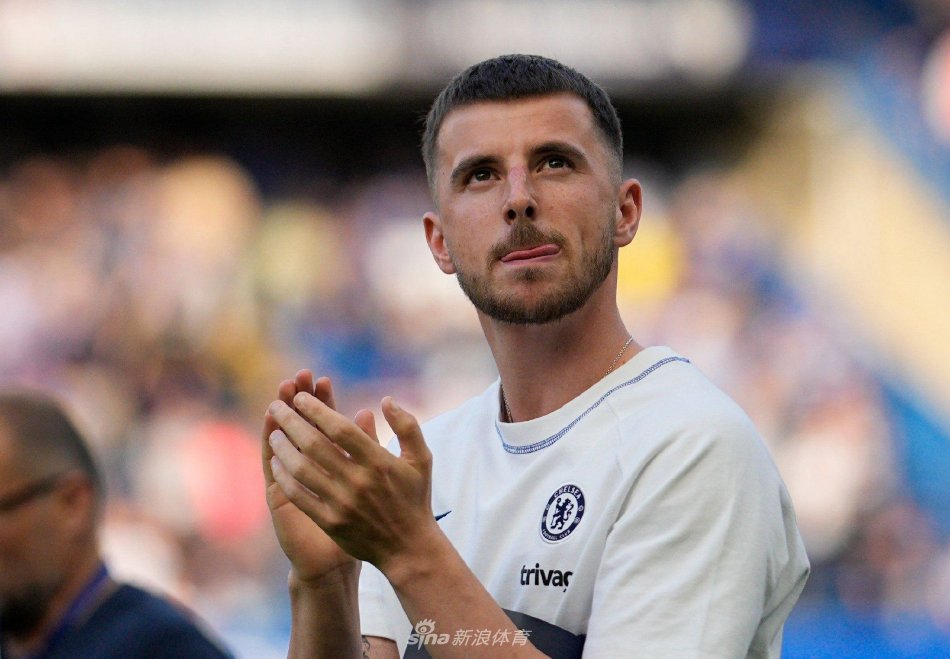 芒特坐在会议桌前，手持签字笔，在镜头前展示签约完成后的喜悦瞬间，背景是曼联俱乐部的标志