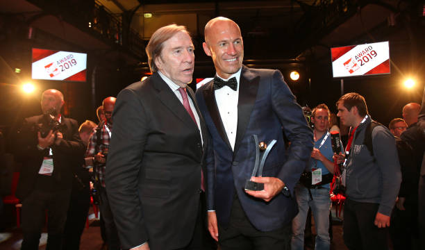 罗本手持奖杯望向远方，眼神深邃而坚定，似乎在回忆那些年用速度撕破防线的瞬间，西装袖口露出精致的腕表