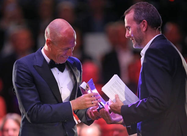 罗本在颁奖台上侧身致意，西装剪裁完美勾勒出运动员的挺拔身姿，背景的闪光灯如同星河般璀璨