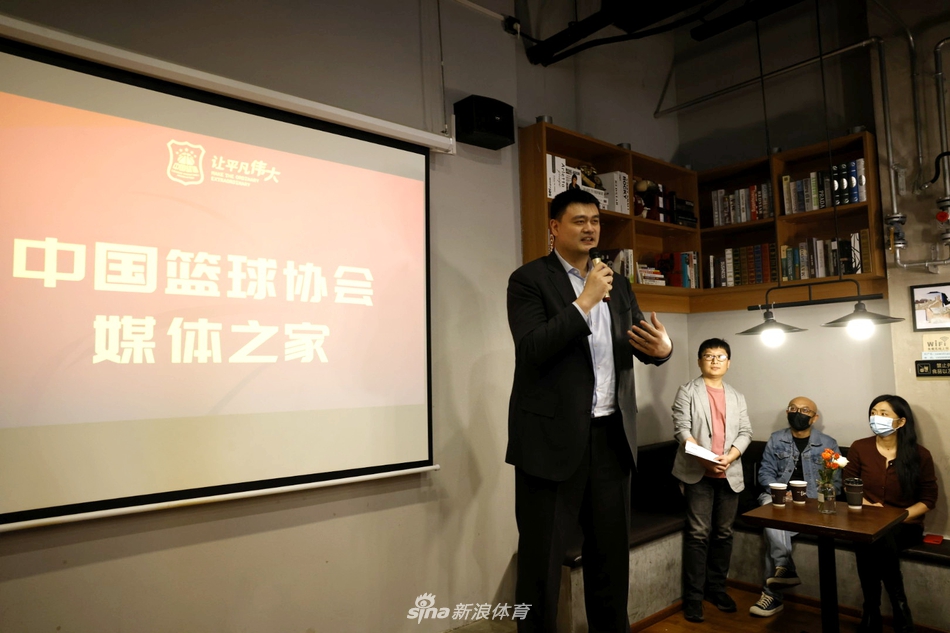 坐在主席台上的姚明，一只手轻轻扶着桌面的文件，侧耳倾听身旁工作人员的发言，嘴角带着一丝若有若无的笑意，目光中流露出对团队工作的信任与支持。