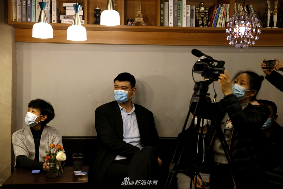 姚明在媒体之家现场，身着深色西装显得干练挺拔，他手持话筒认真倾听记者提问，背景墙上中国篮球协会的标志在灯光下熠熠生辉，整个场景严肃而充满希望。
