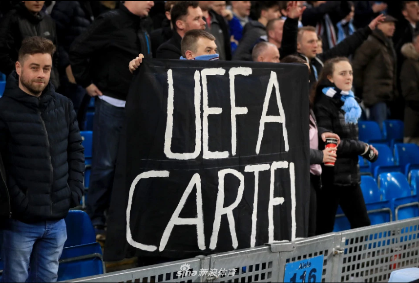 一名年轻的曼城球迷双手举着自制的纸质标语，一张白纸上用黑色记号笔清晰地写着“诈骗犯”的英文单词，他的眼神坚定而专注，身后是座无虚席的看台，这朴素而有力的抗议成为全场愤怒情绪的缩影。