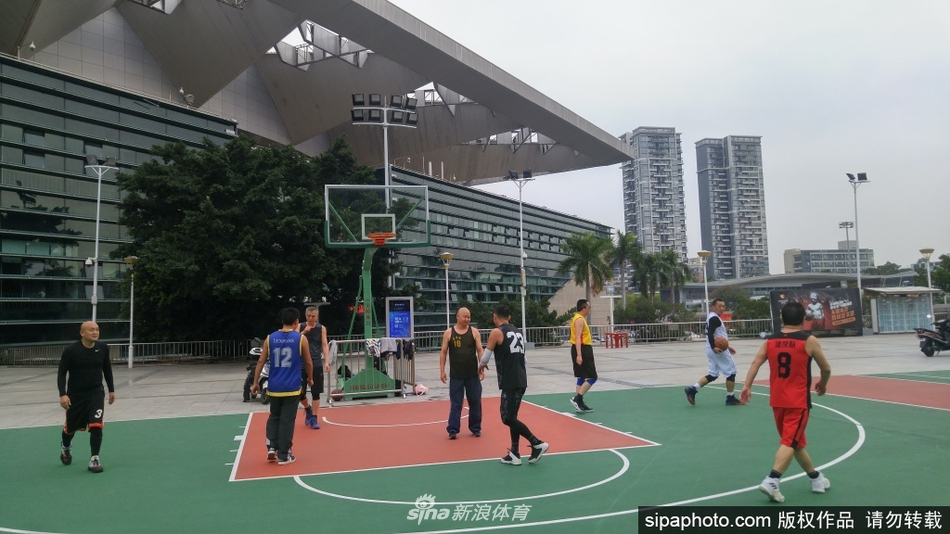 一位身穿白色上衣的男生正在三分线外从容跳投,他的身影在空旷的球场背景下显得格外专注,远处铁丝网外可见城市楼宇,仿佛在静静观看这场久违的运动盛宴