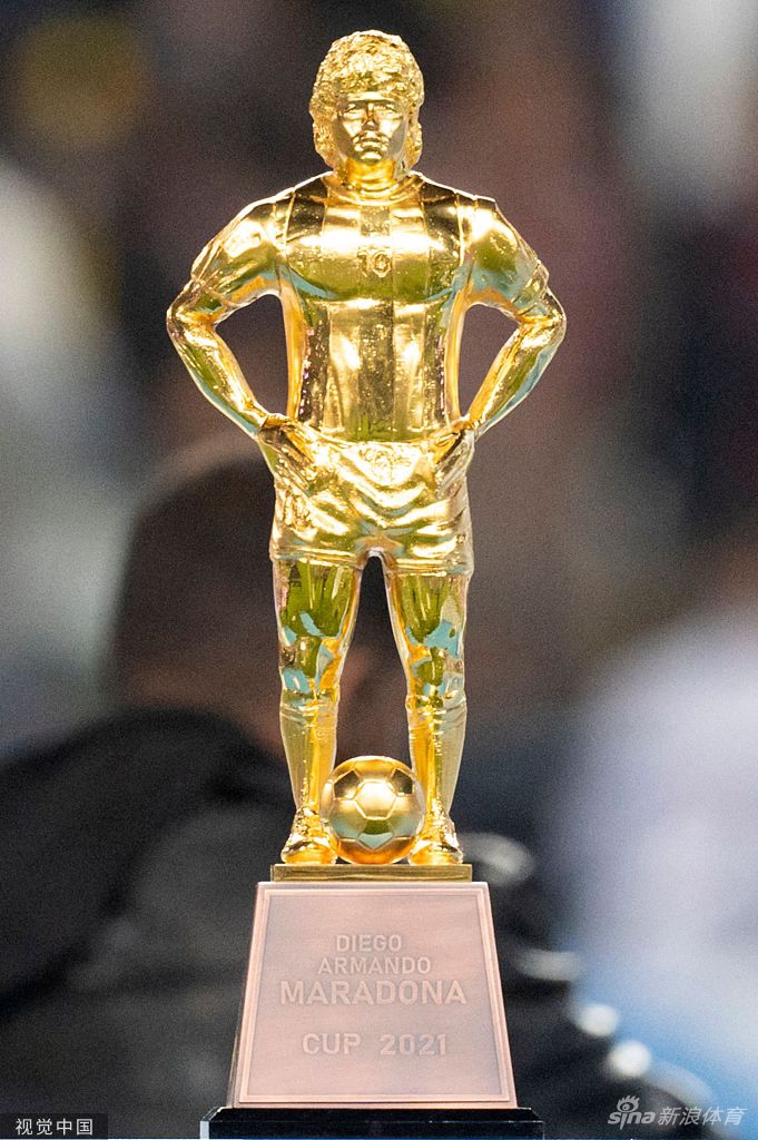 博卡青年队长高举马拉多纳纪念杯，奖杯顶端金光闪耀，全队簇拥在领奖台上振臂高呼，背景是漫天飞舞的彩带。