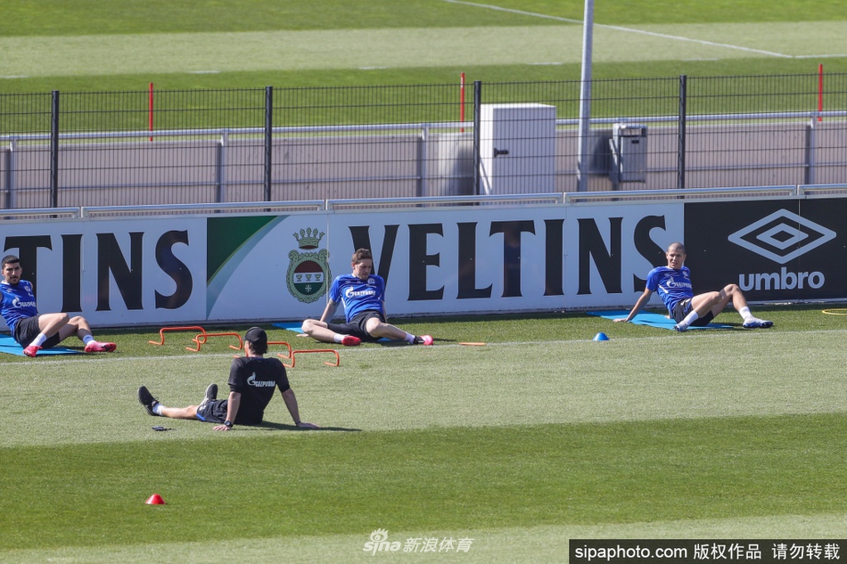 一名沙尔克04球员正在进行带球训练，他的周围空无一人，只有锥筒作为标志。远处，另一名队友正在独自进行拉伸，整个训练场被分割成一个个独立的‘安全岛’。