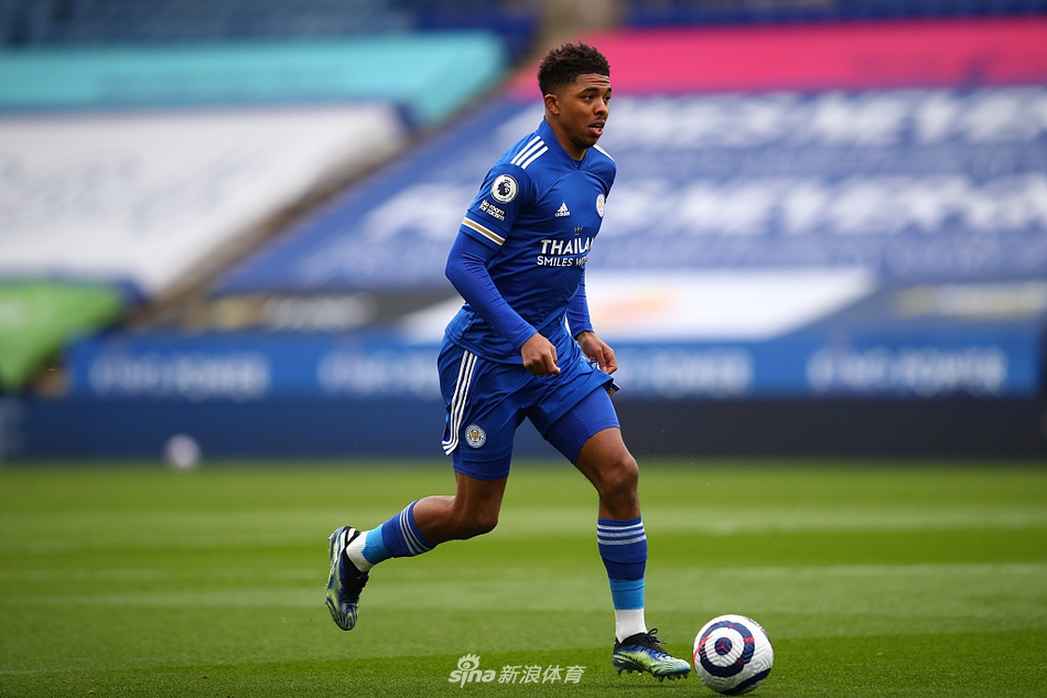 谢菲尔德联门将拉姆斯代尔沮丧地坐在草皮上，叉腰低头，背景是莱斯特城球员庆祝进球的模糊身影