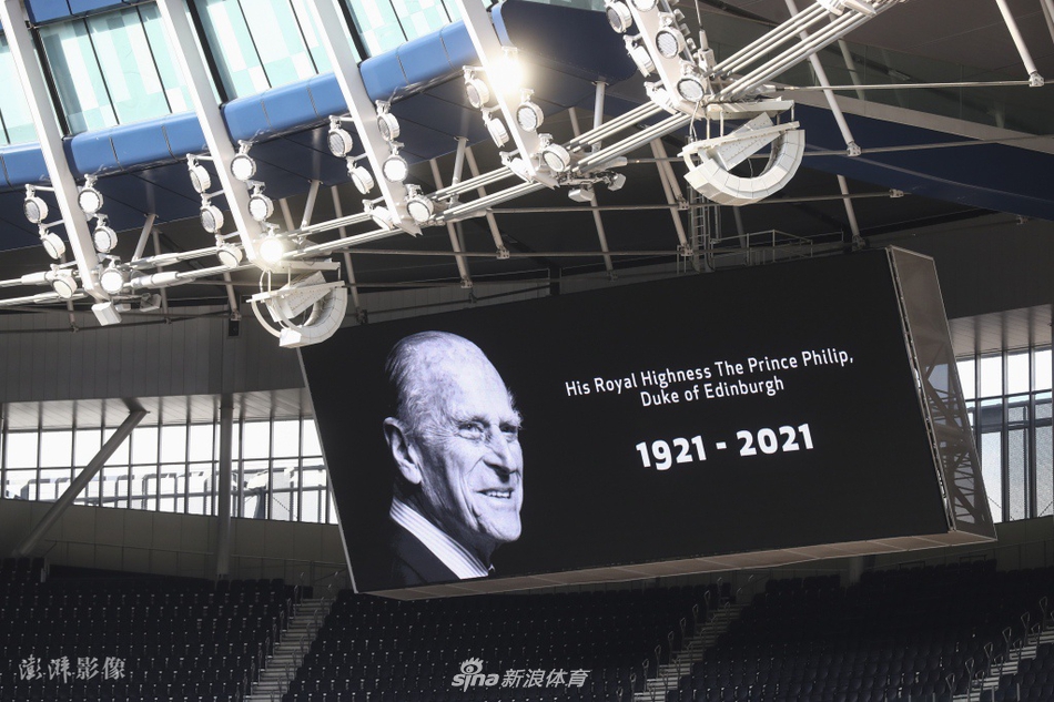 从球场看台高层俯瞰，身穿不同颜色球衣的双方球员与裁判员并肩站在中圈线上，全场寂静无声，共同低头缅怀