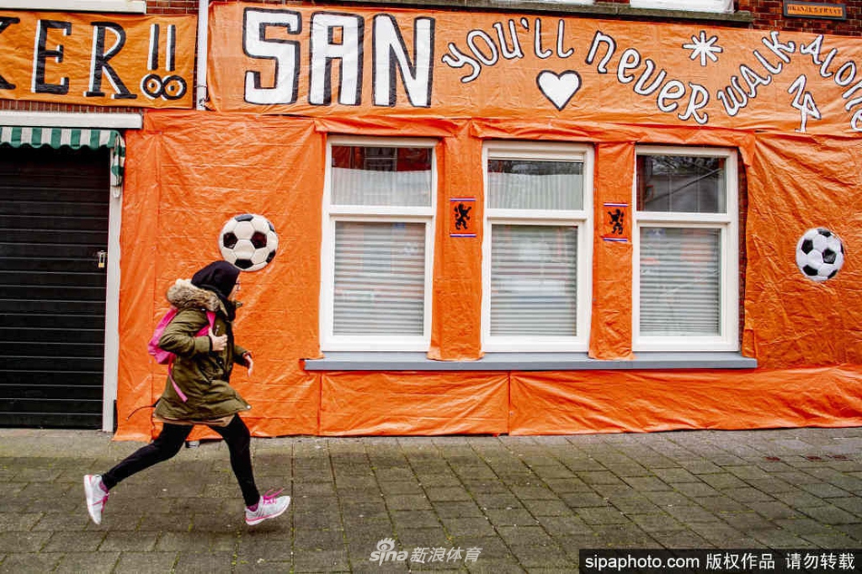 夜幕降临下的海牙市中心广场，大量荷兰球迷聚集在一起观看欧洲杯赛事的大屏幕直播。画面中球迷们手臂相绕，脸上涂着橙白红蓝四色的国旗油彩，正对着屏幕激动地呐喊助威，手中的橙色围巾在灯光下格外醒目，背景是城市建筑上投影出的巨型荷兰国旗图案，场面壮观而热烈。