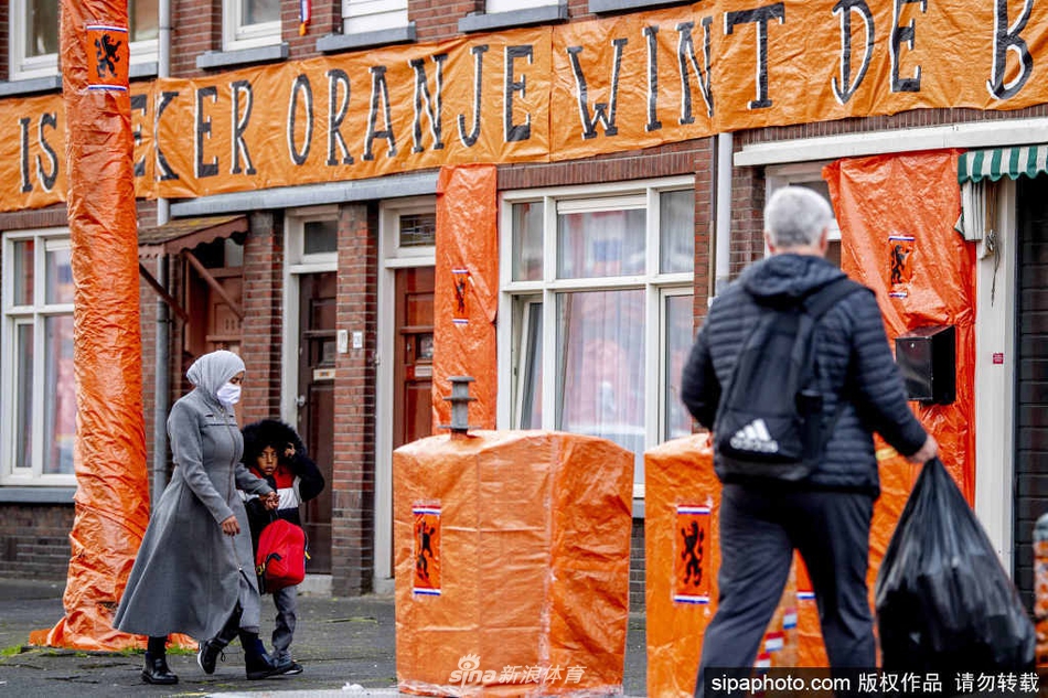 荷兰海牙的一条商业街，街道上空挂满了密密麻麻的荷兰国旗和橙色旗帜，沿街的咖啡馆和商店外也装饰着橙色气球与横幅，三五成群的球迷身着橙色球衣漫步其中，整条街巷仿佛被橙色浸染，洋溢着节日般的欢快气氛。
