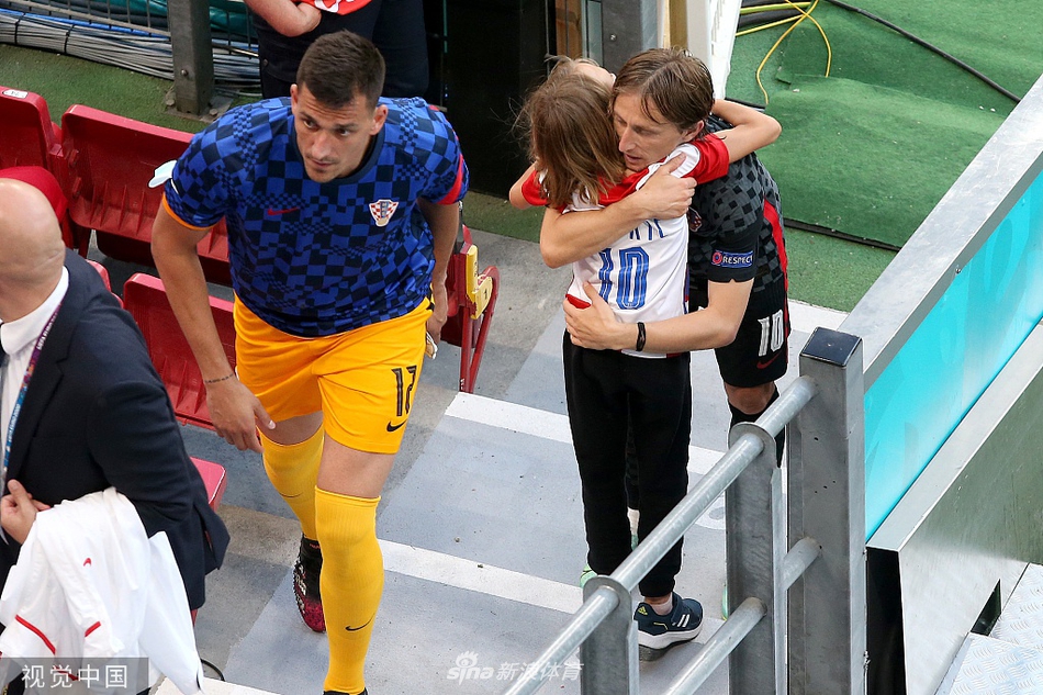 一个穿着克罗地亚球衣的小球迷被父亲抱在怀里，他手里拿着奶嘴，眼神懵懂地望向镜头，身后的球场灯光映衬出这温馨的一幕。